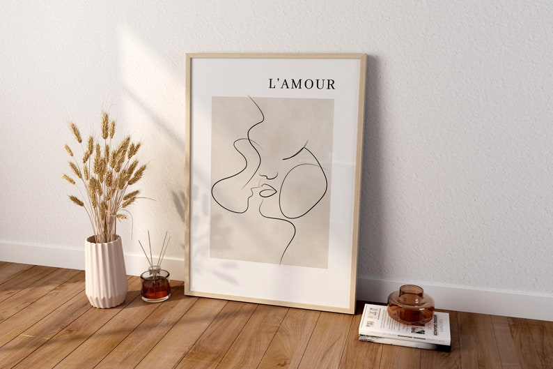May include: A framed print with a minimalist line drawing of two faces, with the text "L'AMOUR" at the top. The print is on a beige background and is framed in a light wood frame. The print is sitting on a wooden floor with a vase of dried wheat and a glass diffuser.