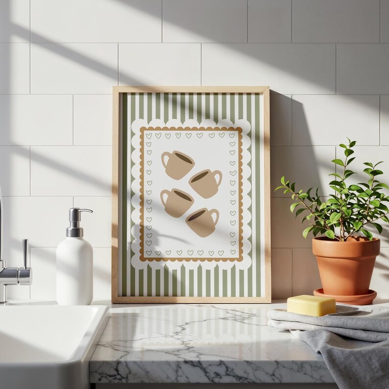May include: A framed print featuring four brown coffee cups on a white background with a scalloped edge and heart accents. The background has green vertical stripes. The frame is light brown. The print is displayed on a countertop with a plant and soap.