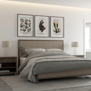 May include: A gray upholstered bed with a headboard featuring a tufted design and nailhead trim. The bed is made up with gray bedding and two white pillows. Three framed art prints hang above the headboard, featuring abstract designs and the text "Galerie d'Art", "Matisse", and "Abstract".