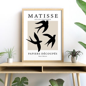 May include: A framed art print featuring a beige background with three black bird silhouettes. The print includes the text "MATISSE AIX-EN-PROVENCE PAPIERS D&Eacute;COUP&Eacute;S Henri Matisse" in black. The artwork is displayed on a light wood table.