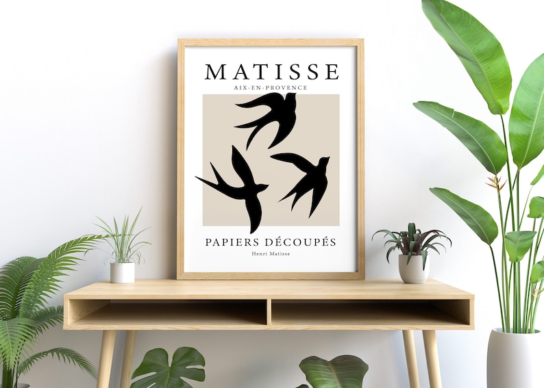 May include: A framed art print featuring a beige background with three black bird silhouettes. The print includes the text "MATISSE AIX-EN-PROVENCE PAPIERS D&Eacute;COUP&Eacute;S Henri Matisse" in black. The artwork is displayed on a light wood table.