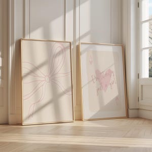 May include: Two framed art prints. One features a light pink bow design on a beige background. The other print has the text "I'm literally just a girl" with a pink heart on a beige background. Both are in gold frames.