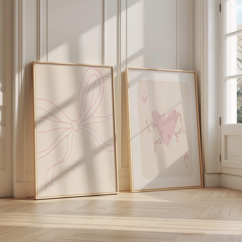 May include: Two framed art prints. One features a light pink bow design on a beige background. The other print has the text "I'm literally just a girl" with a pink heart on a beige background. Both are in gold frames.