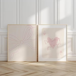 May include: Two framed art prints. One features a light pink bow design on a beige background. The other print has a pink heart and the text "I'm literally just a girl" on a beige background. Both are in gold frames.