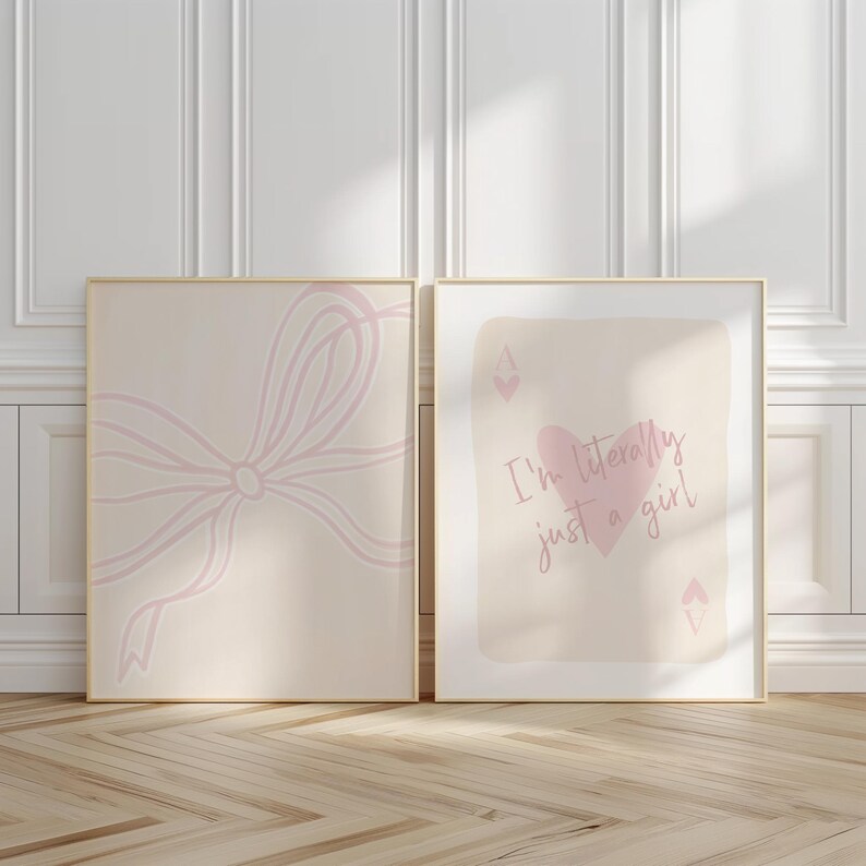 May include: Two framed art prints. One features a light pink bow design on a beige background. The other print has a pink heart and the text "I'm literally just a girl" on a beige background. Both are in gold frames.