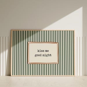 Puede incluir: Arte de pared enmarcado con un fondo a rayas verdes y blancas. La obra de arte presenta una secci&oacute;n rectangular de color crema con las palabras "kiss me good night" en negro, en una fuente de estilo m&aacute;quina de escribir. El marco es de color madera clara.