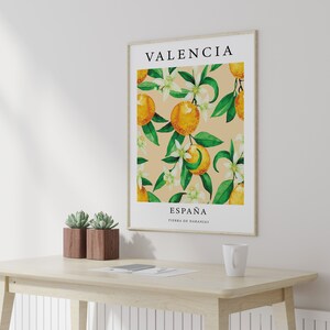 Op de afbeelding: Een ingelijste poster met een aquarel-illustratie van sinaasappelbloesems en sinaasappelen. De poster heeft de tekst "Valencia, Espa&ntilde;a, Tierra de Naranjas" erop gedrukt.