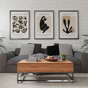 May include: Three framed black and white art prints with beige backgrounds hanging on a white brick wall above a gray sofa with gray pillows. The prints feature abstract designs and the text "Galerie d'Art", "Matisse", and "Abstract".