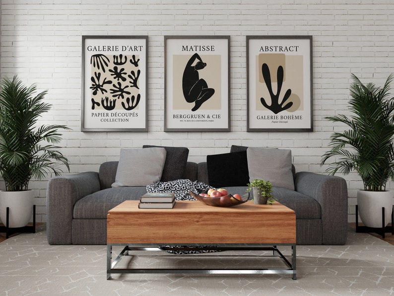 May include: Three framed black and white art prints with beige backgrounds hanging on a white brick wall above a gray sofa with gray pillows. The prints feature abstract designs and the text "Galerie d'Art", "Matisse", and "Abstract".