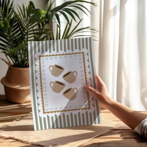 May include: A framed print featuring four coffee cups in a light brown color, arranged on a white background with a decorative border of hearts and a scalloped edge. The print has a green and white striped border.