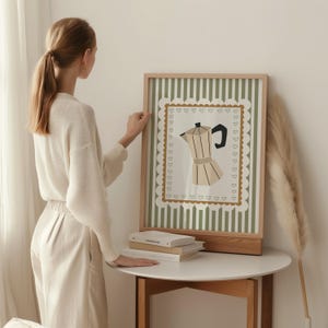 Peut inclure: Une affiche encadr&eacute;e avec une illustration de cafeti&egrave;re. L'&oelig;uvre d'art a un cadre en bois brun clair et un fond &agrave; rayures vertes et blanches. La cafeti&egrave;re est beige avec une poign&eacute;e noire. L'affiche est pos&eacute;e sur une table blanche.