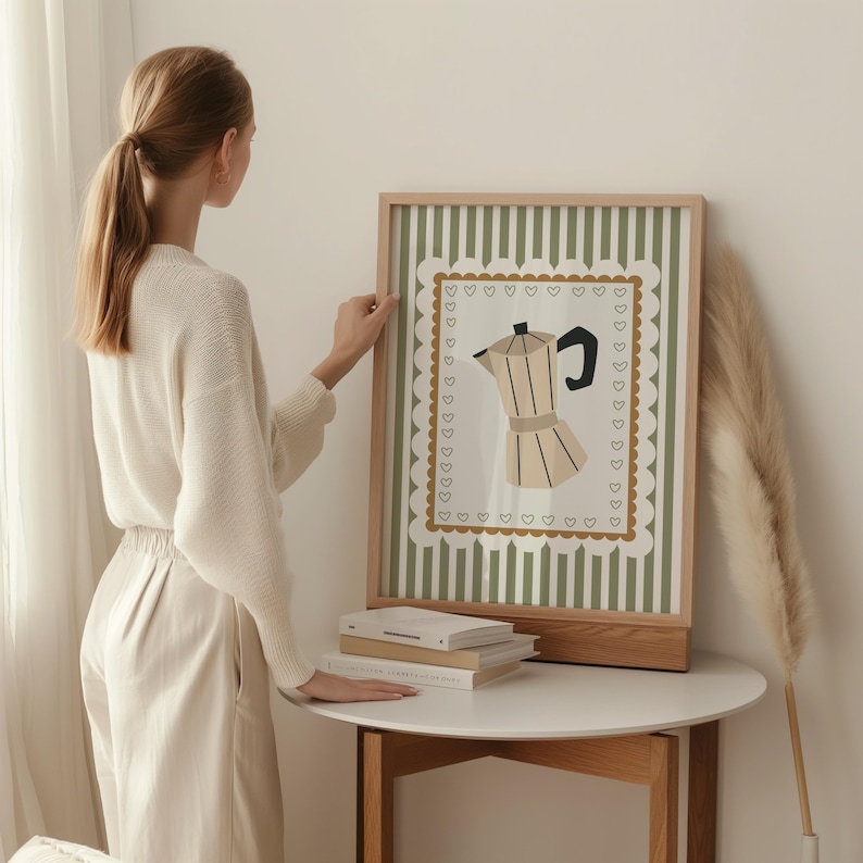 Peut inclure: Une affiche encadr&eacute;e avec une illustration de cafeti&egrave;re. L'&oelig;uvre d'art a un cadre en bois brun clair et un fond &agrave; rayures vertes et blanches. La cafeti&egrave;re est beige avec une poign&eacute;e noire. L'affiche est pos&eacute;e sur une table blanche.