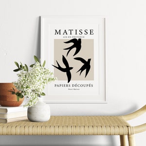 May include: A framed art print featuring a beige background with three black bird silhouettes. The print includes the text "MATISSE AIX-EN-PROVENCE PAPIERS D&Eacute;COUP&Eacute;S" in black. The artwork is displayed on a light-colored bench with a vase of white flowers.