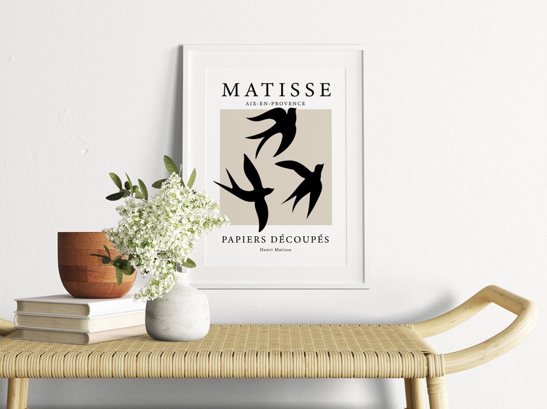 May include: A framed art print featuring a beige background with three black bird silhouettes. The print includes the text "MATISSE AIX-EN-PROVENCE PAPIERS D&Eacute;COUP&Eacute;S" in black. The artwork is displayed on a light-colored bench with a vase of white flowers.