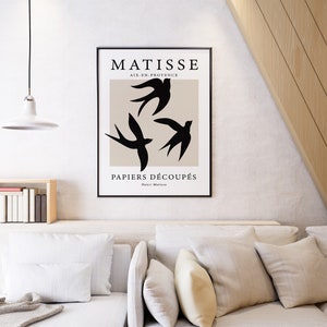 May include: A framed art print featuring a minimalist design with three black bird silhouettes against a beige background. The print includes the text "MATISSE AIX-EN-PROVENCE PAPIERS D&Eacute;COUP&Eacute;S" in black font. The artwork is displayed in a modern living room setting.