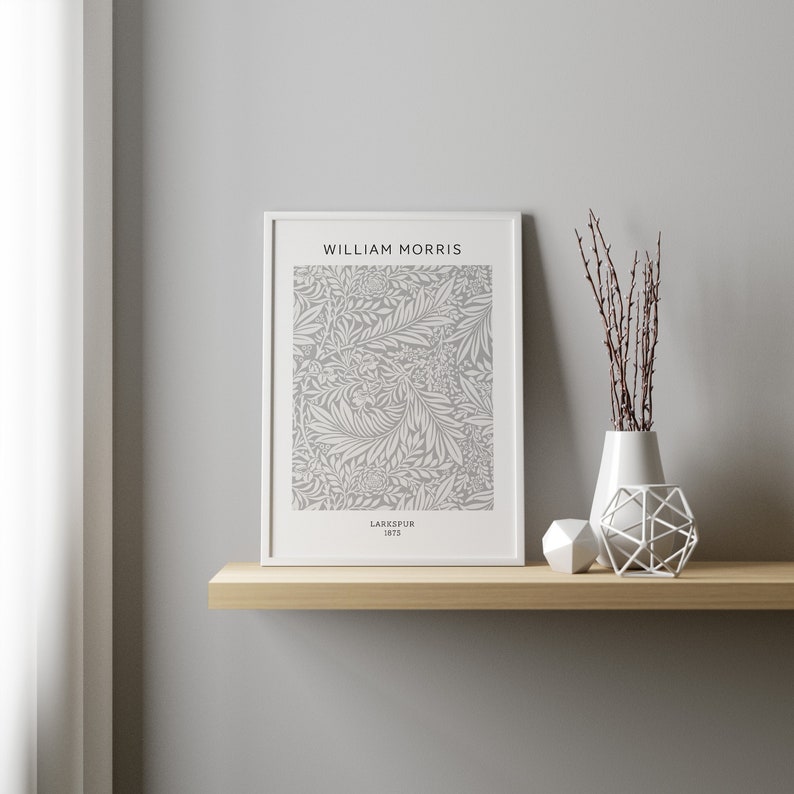 May include: A framed print featuring a gray and white floral pattern, with the text "WILLIAM MORRIS" and "LARKSPUR 1875". The print is displayed on a wooden shelf, alongside a white vase with branches, and two geometric decorative objects.