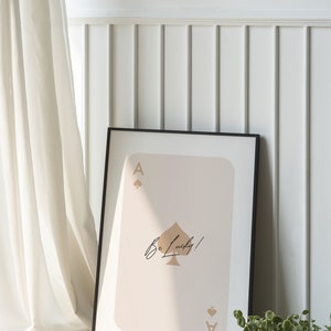 May include: A black framed print of an ace of spades playing card with a beige background. The ace of spades is gold and the text "Be Lucky!" is written in black cursive.