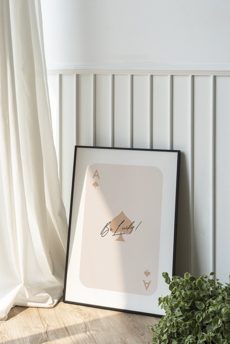 May include: A black framed print of an ace of spades playing card with a beige background. The ace of spades is gold and the text "Be Lucky!" is written in black cursive.