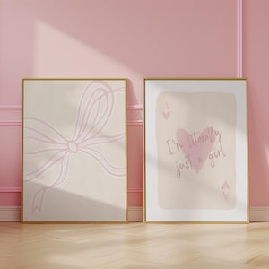 May include: Two framed art prints with gold frames. One features a light pink bow on a beige background. The other has a playing card design with a pink heart and the text "I'm literally just a girl."