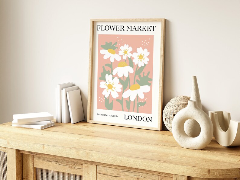 May include: A framed print with a pink background and white daisies with yellow centers. The print says "FLOWER MARKET" at the top and "THE FLORAL GALLERY LONDON" at the bottom.