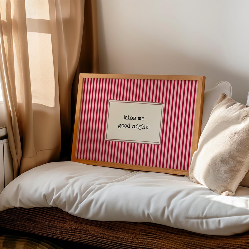 Puede incluir: Una obra de arte enmarcada con un marco de madera. La obra presenta un fondo a rayas rojas y blancas con una inserci&oacute;n rectangular con el texto "kiss me good night". La obra est&aacute; colocada sobre una almohada blanca.