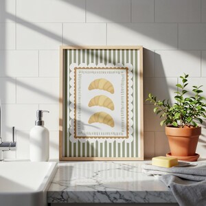 May include: A framed print of three golden croissants. The artwork has a green and white striped background with a scalloped border. The frame is light brown, and the print is displayed on a countertop.