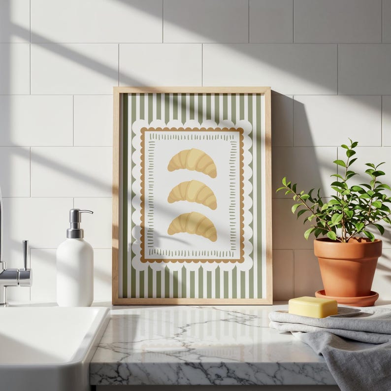 May include: A framed print of three golden croissants. The artwork has a green and white striped background with a scalloped border. The frame is light brown, and the print is displayed on a countertop.