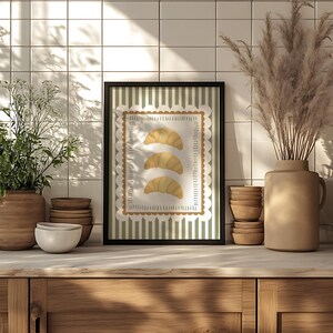 May include: A framed print depicting three croissant illustrations. The artwork has a white scalloped border and a green and white striped background. The print is displayed on a wooden surface, alongside decorative items.