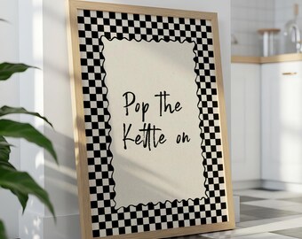 Op de afbeelding: Een ingelijste print met de tekst "Pop the Kettle on". Het kunstwerk heeft een crèmekleurige achtergrond, zwarte tekst en een zwart-wit geblokte rand. De lijst is gemaakt van lichtgekleurd hout.