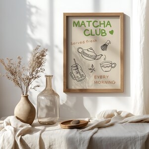 Pu&ograve; includere: Una stampa incorniciata con le parole "Matcha Club" in verde, insieme a illustrazioni di una teiera, una frusta e una tazza. &Egrave; incluso anche il testo "Served fresh" e "Every morning". La cornice &egrave; di colore marrone chiaro.