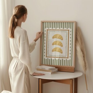 May include: A framed art print with three yellow croissants on a white background, framed by a scalloped border. The background has green vertical stripes. The frame is made of light-coloured wood.