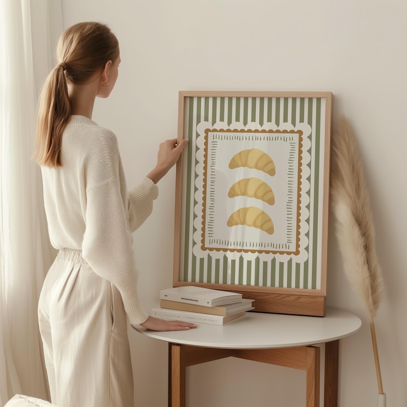 May include: A framed art print with three yellow croissants on a white background, framed by a scalloped border. The background has green vertical stripes. The frame is made of light-coloured wood.