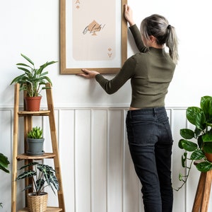 May include: A person hangs a framed print of an ace of spades playing card with the text "Be Lucky!" on a white wall. The print has a light pink background and brown lettering. The person is wearing a green sweater and black jeans.
