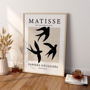 May include: A framed art print featuring a minimalist design with three black swallows against a beige backdrop. The print includes the text "MATISSE AIX-EN-PROVENCE PAPIERS D&Eacute;COUP&Eacute;S Henri Matisse". The artwork is displayed on a wooden floor.