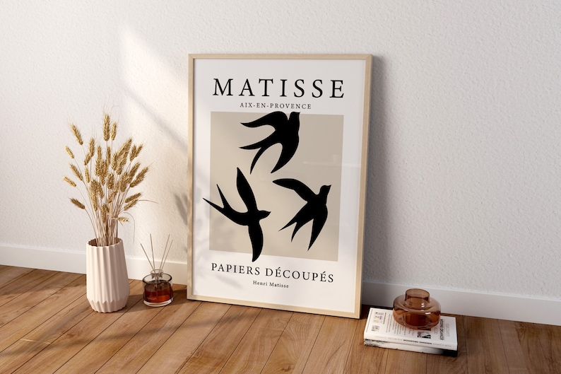 May include: A framed art print featuring a minimalist design with three black swallows against a beige backdrop. The print includes the text "MATISSE AIX-EN-PROVENCE PAPIERS D&Eacute;COUP&Eacute;S Henri Matisse". The artwork is displayed on a wooden floor.