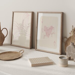 May include: Two framed art prints with light wood frames. One print features an abstract floral design in pink and beige. The other print has a pink heart and the text "I'm literally just a girl".