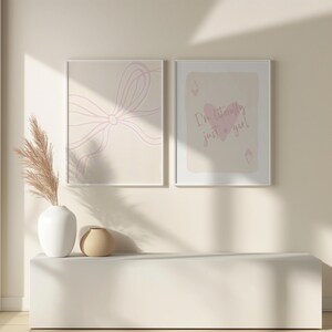 May include: Two framed art prints. One features a light pink bow design on a beige background. The other print has a playing card design with the text "I'm literally just a girl" in pink. A white vase with dried plants and a small beige vase are on a white shelf.
