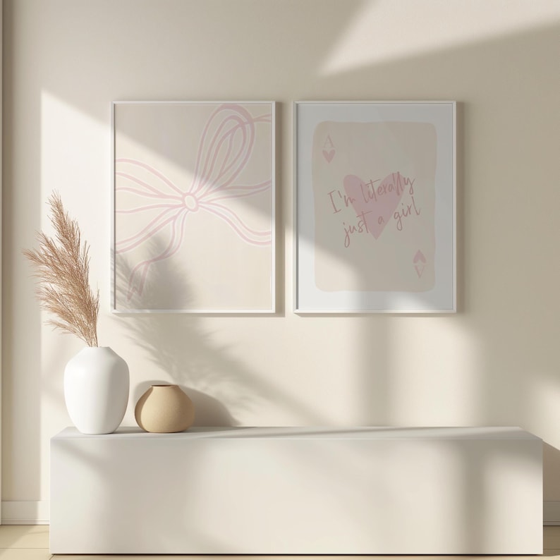May include: Two framed art prints. One features a light pink bow design on a beige background. The other print has a playing card design with the text "I'm literally just a girl" in pink. A white vase with dried plants and a small beige vase are on a white shelf.