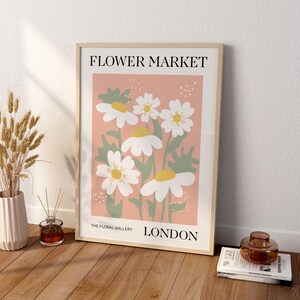 May include: A framed poster with a pink background and a floral illustration of white daisies with yellow centers. The poster reads "FLOWER MARKET" at the top, "THE FLORAL GALLERY" at the bottom left, and "LONDON" at the bottom right.