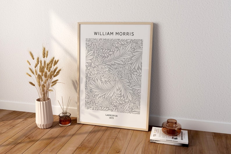 May include: A framed print featuring a floral pattern in gray and white, with the text "WILLIAM MORRIS" at the top and "LARKSPUR 1875" at the bottom. The print is displayed on a wooden floor, next to a vase with wheat stalks.