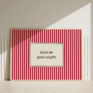 Puede incluir: Un estampado enmarcado con un fondo a rayas rojas y blancas. El estampado presenta un inserto rectangular de color crema con las palabras "kiss me good night" en una fuente negra simple. El marco es de color madera clara.