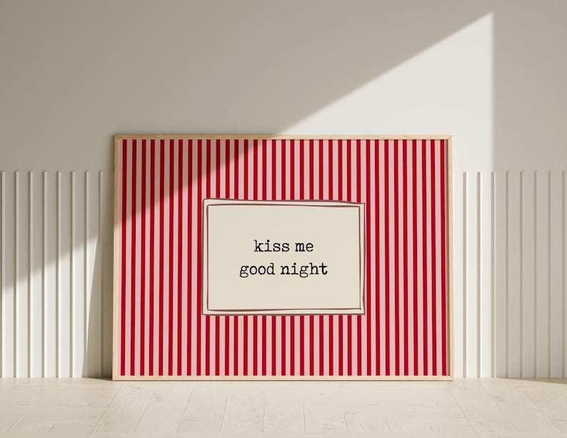 Puede incluir: Un estampado enmarcado con un fondo a rayas rojas y blancas. El estampado presenta un inserto rectangular de color crema con las palabras "kiss me good night" en una fuente negra simple. El marco es de color madera clara.