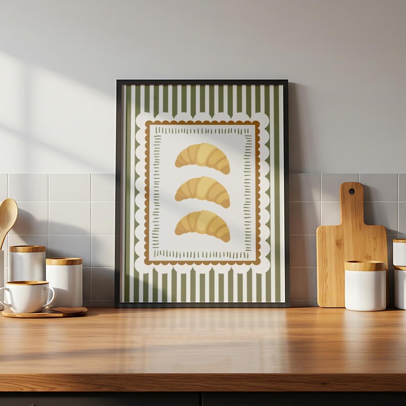 May include: A framed art print showing three golden croissants on a white background with a scalloped border. The background has vertical green and white stripes. The print is displayed on a wooden surface.