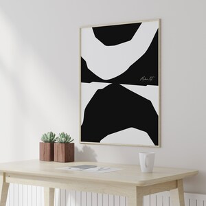 Op de afbeelding: Een zwart-witte abstracte kunstprint in een houten lijst die aan een witte muur hangt boven een houten tafel met twee potplanten met vetplanten. De print heeft een eenvoudig ontwerp van overlappende zwarte en witte vormen. De handtekening van de kunstenaar, "Modern Art", is zichtbaar in de rechterbenedenhoek van de print.