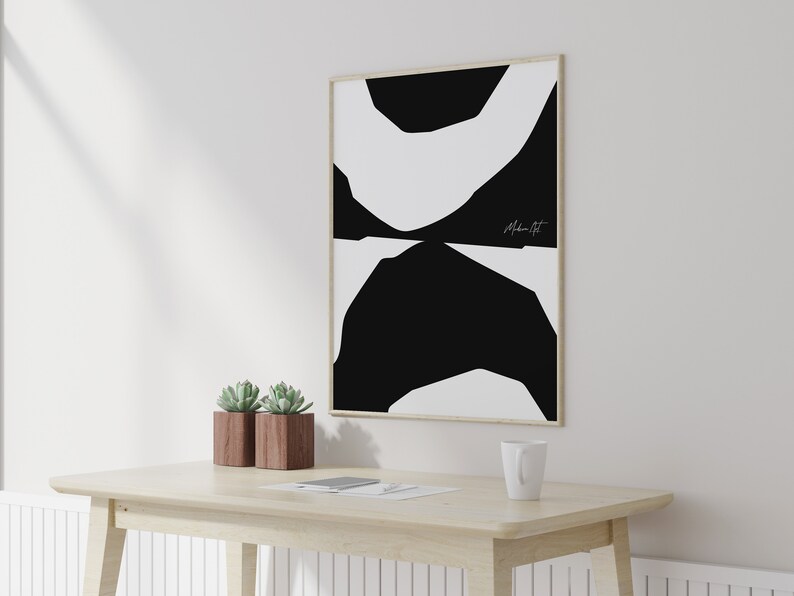 Op de afbeelding: Een zwart-witte abstracte kunstprint in een houten lijst die aan een witte muur hangt boven een houten tafel met twee potplanten met vetplanten. De print heeft een eenvoudig ontwerp van overlappende zwarte en witte vormen. De handtekening van de kunstenaar, "Modern Art", is zichtbaar in de rechterbenedenhoek van de print.