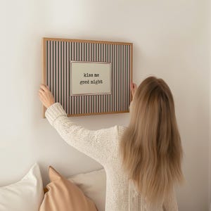 Puede incluir: Un cuadro enmarcado con un fondo a rayas negras y rosas, y un panel rectangular central con el texto "kiss me good night". El marco es de madera.