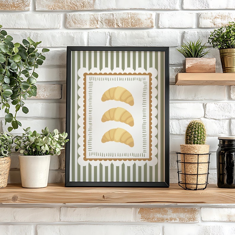 May include: A framed art print showing three golden croissants on a white background with a scalloped border. The background has vertical green stripes. The black framed print is displayed on a wooden shelf.