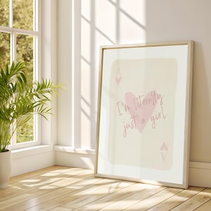 May include: A framed print with the text "I'm literally just a girl" and a pink heart and spade symbols. The artwork is set against a light background, leaning against a white wall near a window and a potted plant.