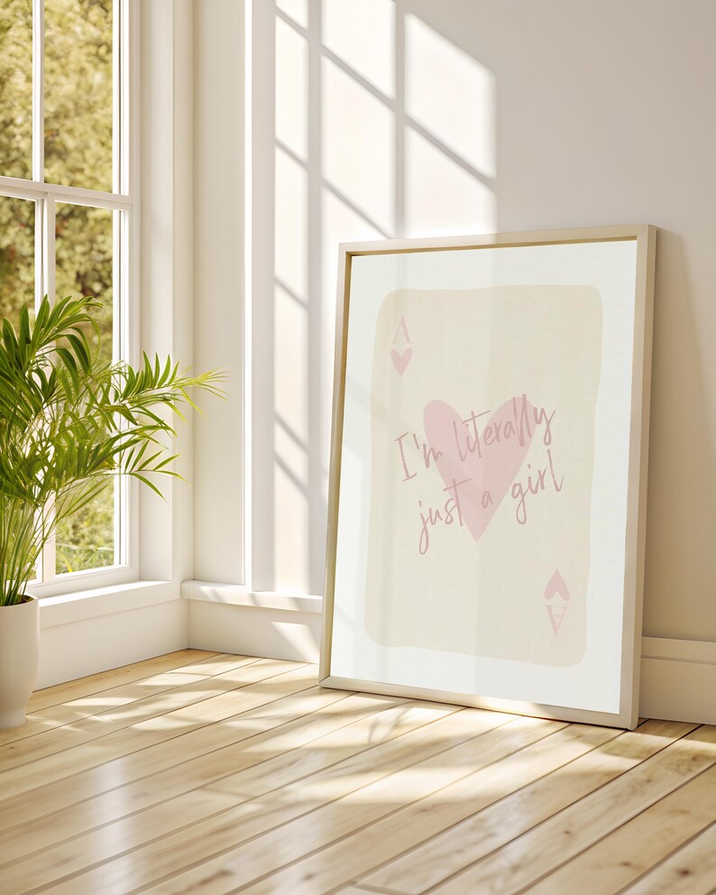 May include: A framed print with the text "I'm literally just a girl" and a pink heart and spade symbols. The artwork is set against a light background, leaning against a white wall near a window and a potted plant.