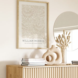 May include: A framed print of a beige floral pattern with the text "WILLIAM MORRIS THE VICTORIA AND ALBERT MUSEUM" at the bottom. The print is hanging on a white wall above a wooden cabinet with a round mirror and a vase with dried flowers.